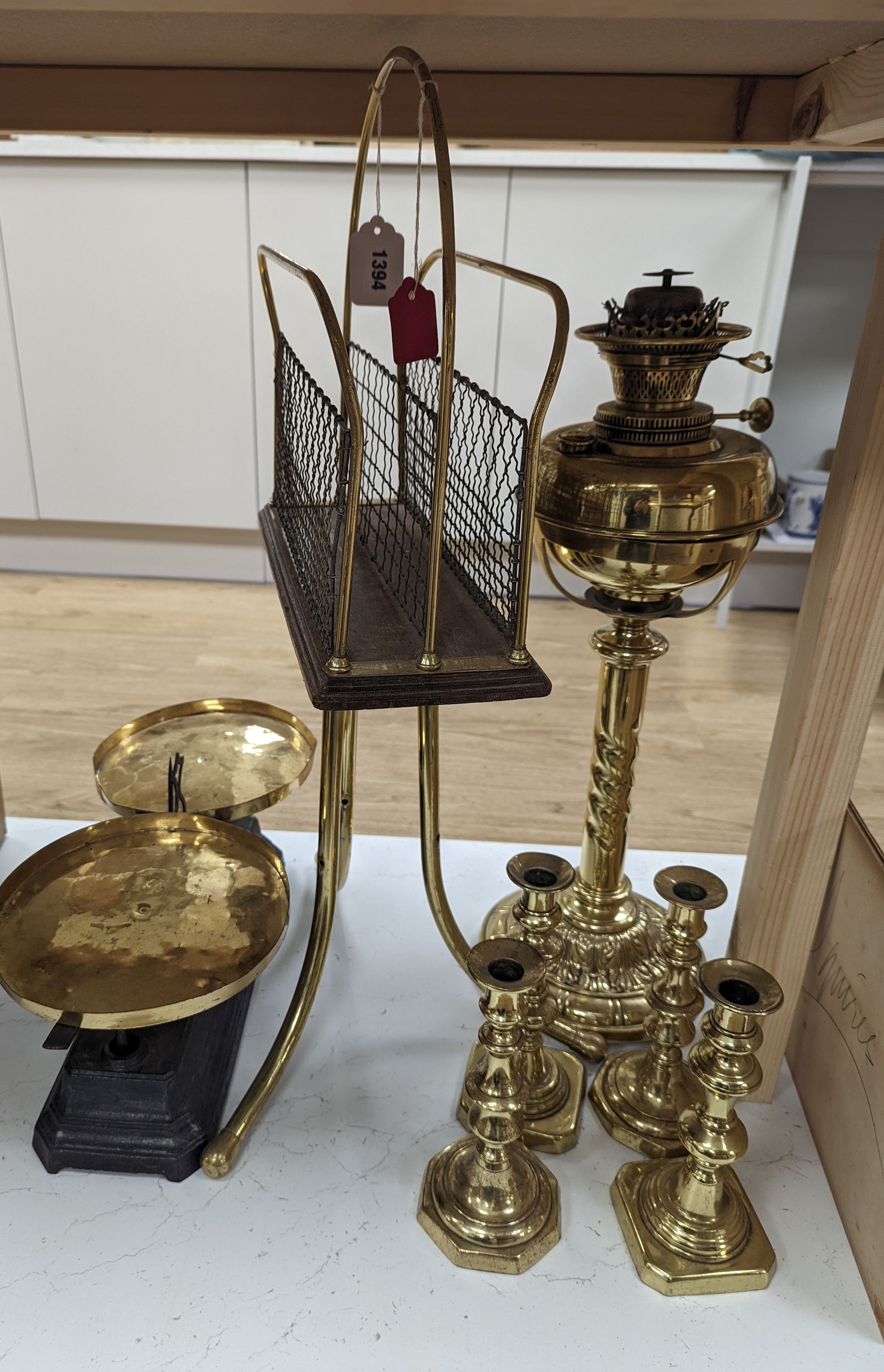 A brass magazine stand, a pair of scales, four candlesticks and a tall brass oil lamp base, Oil lamp 58 cms high.